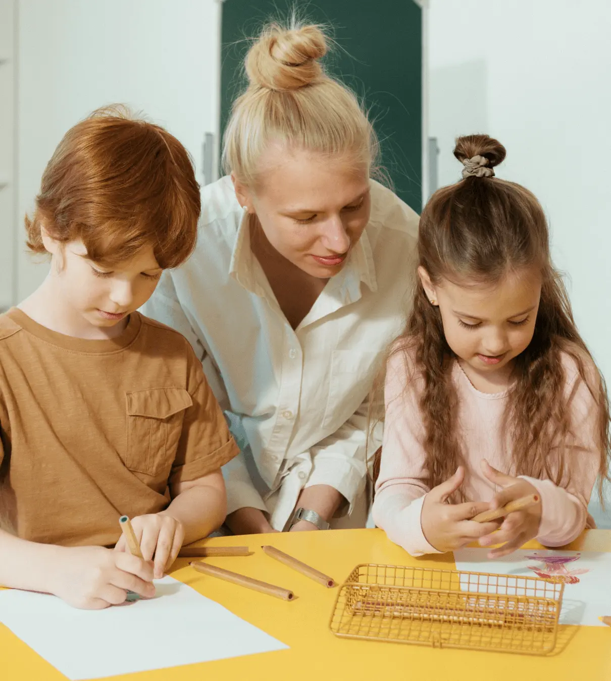 Two students use color pencils to draw on white paper while an educator looks on.